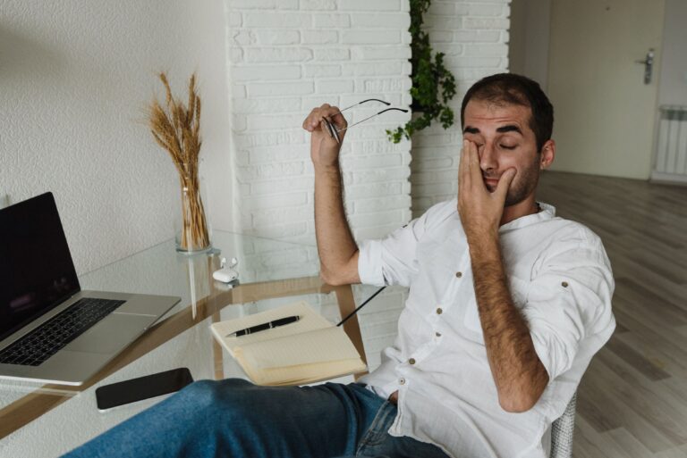 Hombre en home office frotándose el ojo con gafas en la mano, representando fatiga visual por uso de pantallas