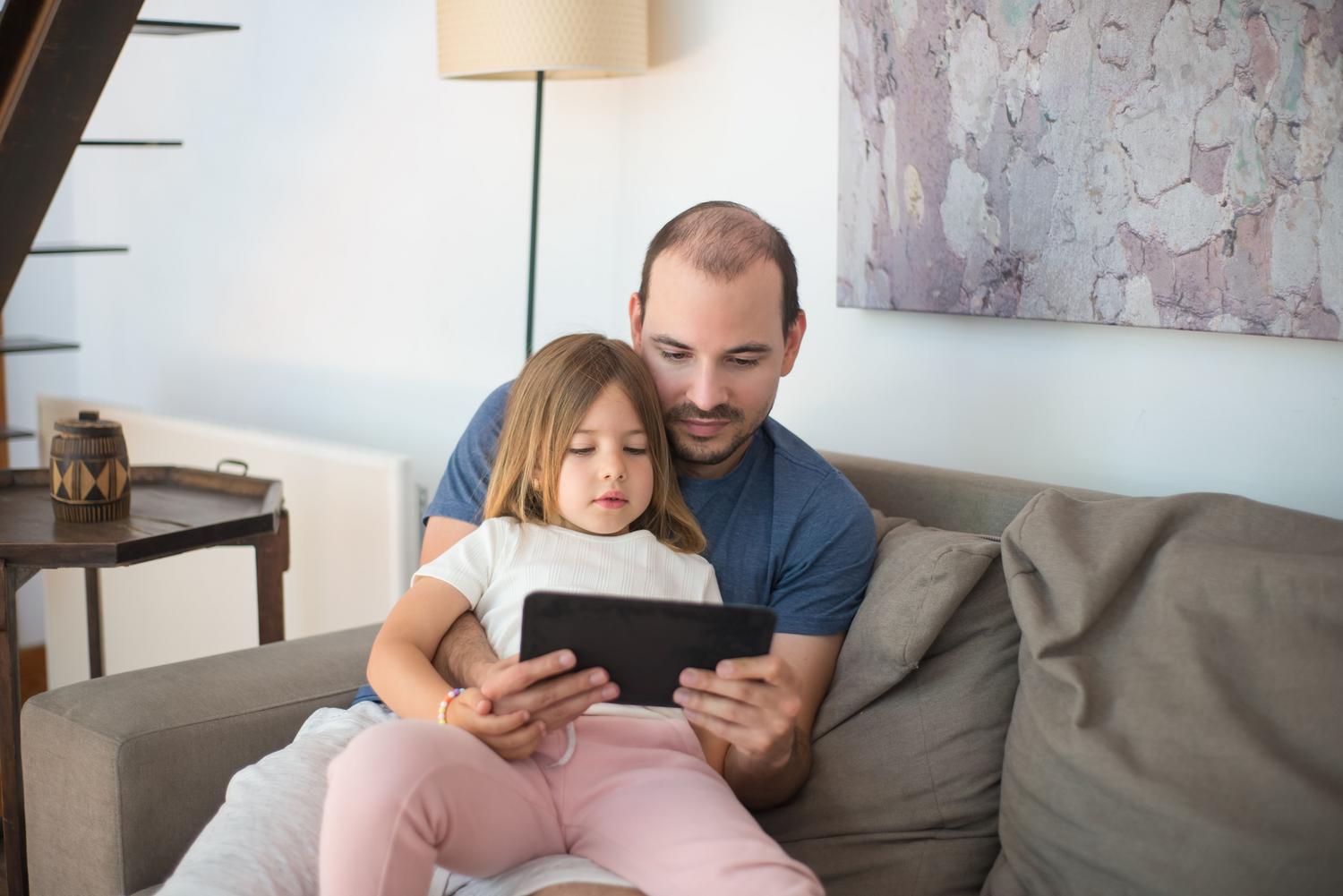 Padre y hija compartiendo el uso de una tablet en el sofá, simbolizando la confianza y la guía parental. Padre y hija compartiendo el uso de una tablet en el sofá, simbolizando la confianza y la guía parental.