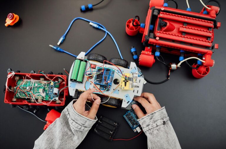 Manos de un entusiasta Maker ensamblando un coche robot con chasis blanco, cables de colores y placa de microcontrolador sobre un fondo oscuro.