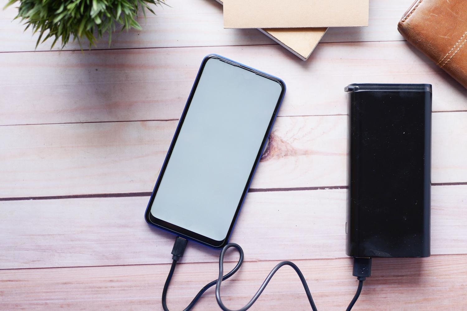 Smartphone cargando con una batería externa sobre un escritorio de madera, representando el uso cotidiano de la energía portátil. Smartphone cargando con una batería externa sobre un escritorio de madera, representando el uso cotidiano de la energía portátil.